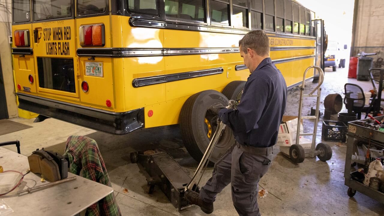 Tire service technician works on a heavy-duty wheel in a shop, with an α logo on the coach.